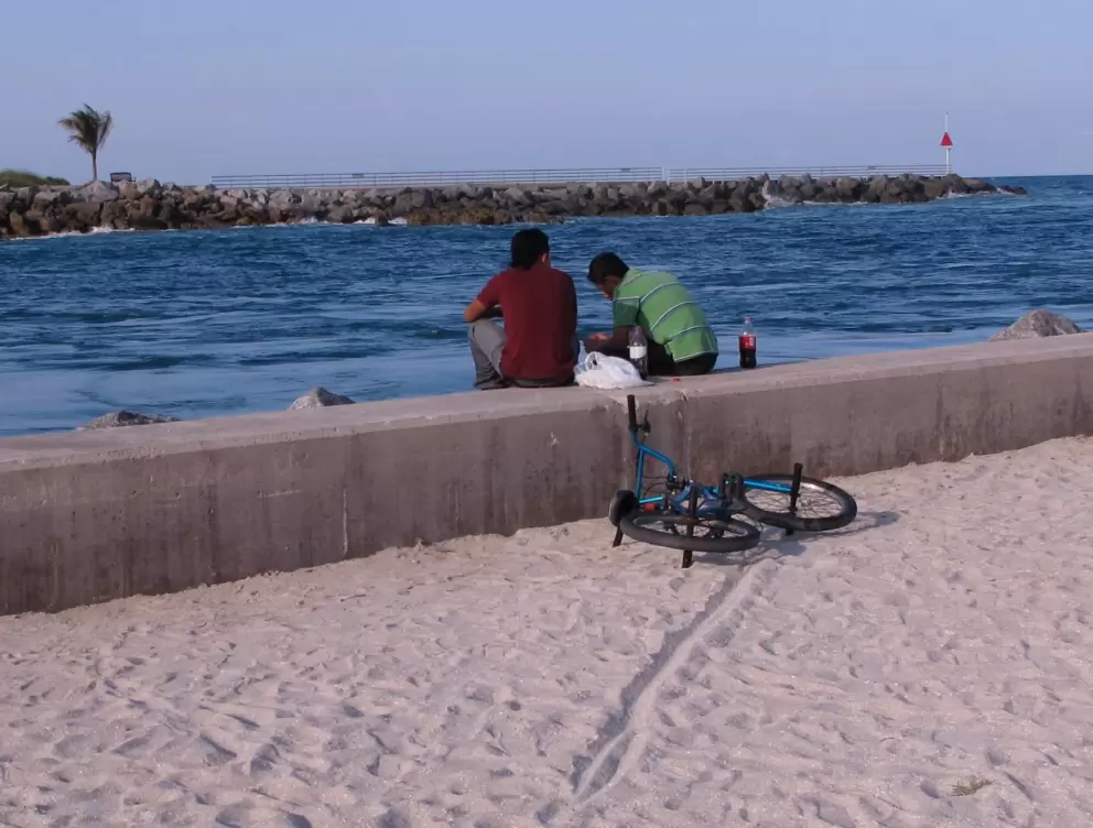 Two boys sit on the cement sea wall, bike in the sand.
