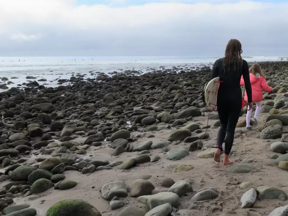 A surfer girl walks along the beach as the sun tries to break through.