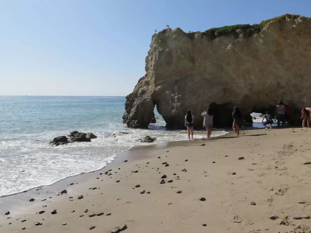 Beachgoers watch water rush under an arch.