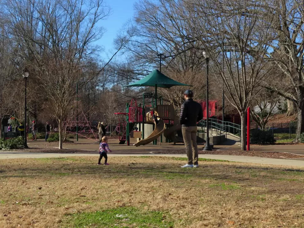 A dad stands with his toddler on the grass in the middle of the play area.