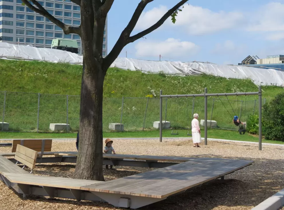 Wooden bench around the base of a tree, plus swings at Esplanade Park. Work on the fortifications in the background. 