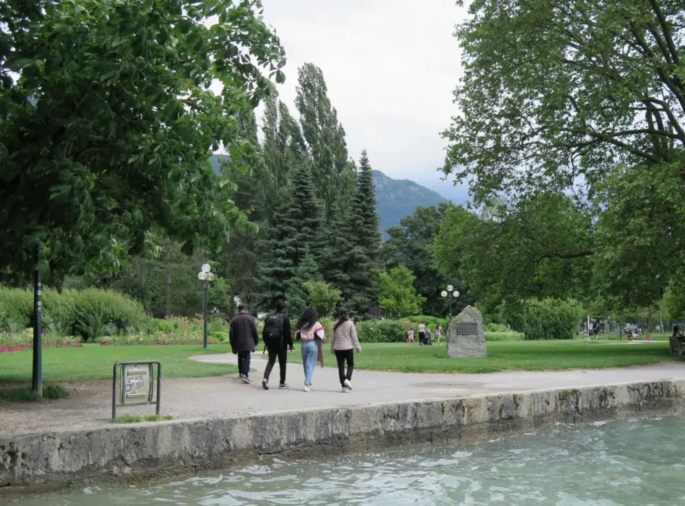 Friends walking among pine trees, lamposts, and flowers at Parc Charles Bosson. 
