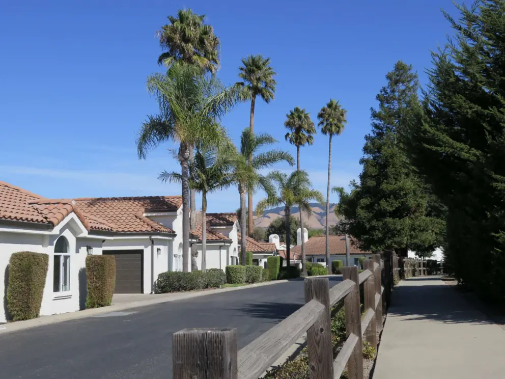 Low wooden fence between Country Club Dr and Pebble Beach Way. Walkway with some shade, and views of palms and Spanish tiled roofs.