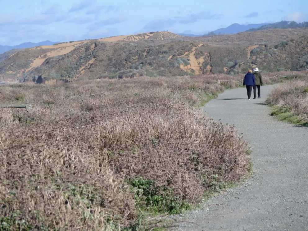 A couple walks the trail, with red dirt mountain in the distance.