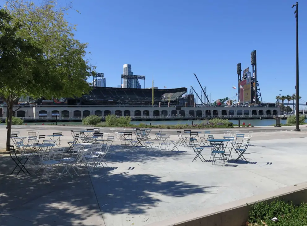 Chairs and tables at China Basin Park, with Oracle Park Stadium across the water. 