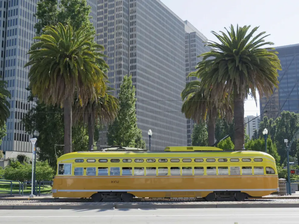 A yellow and orange historic trolley, at Embarcadero, with canary palms behind.