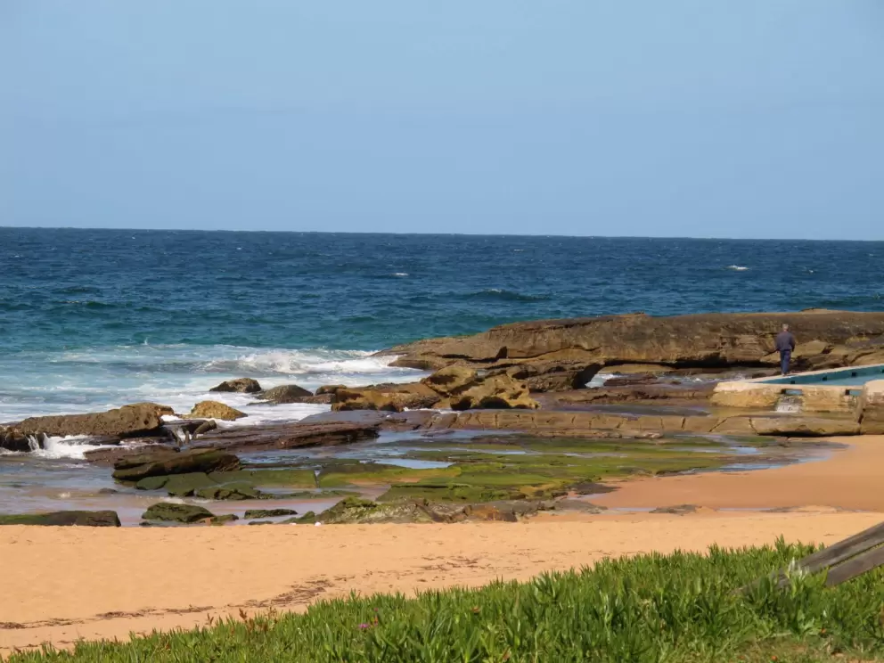 Man in jacket enjoying the rocky end of the beach by the ocean swimming pool.