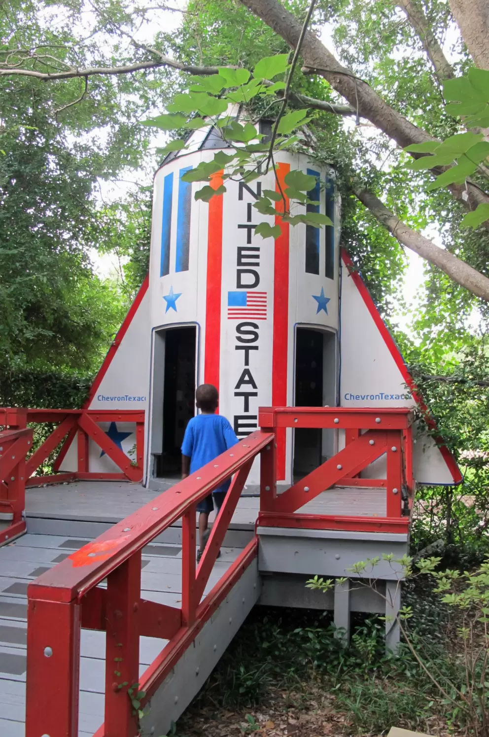 A boy checks out the rocket ship under the leafy trees.