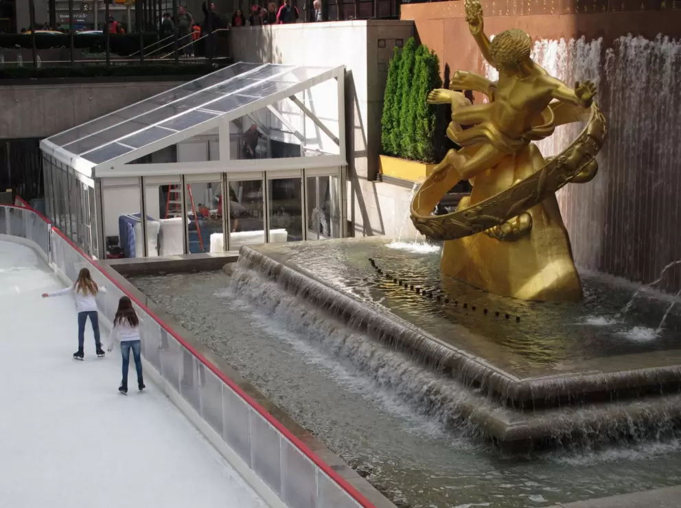 Two girls ice skate past the golden statue.