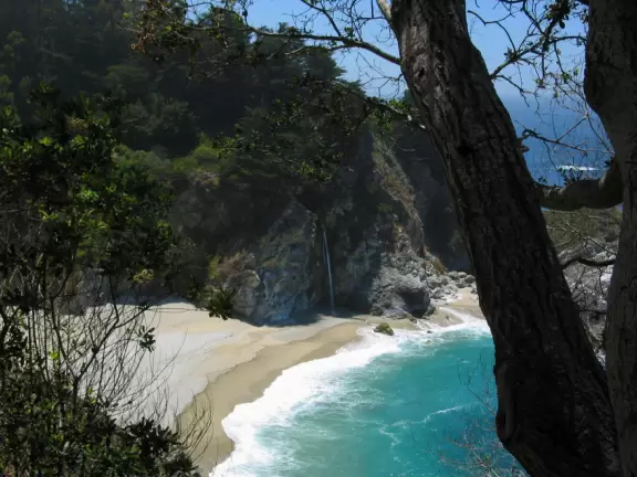 A stunning hike overlooking a white sand beach, bright blue-green waters, and a waterfall falling onto the beach.